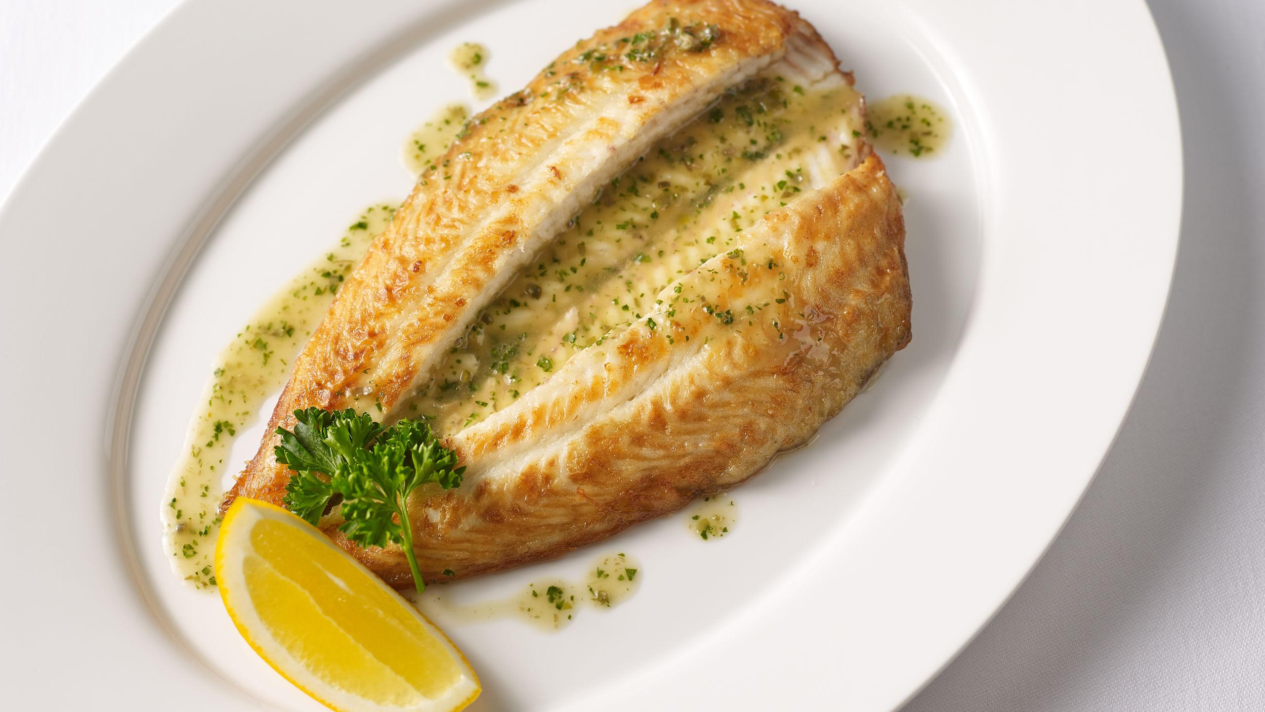 Whole pan-seared Dover sole on a white oval plate with lemon butter sauce, capers, parsley, and a lemon wedge