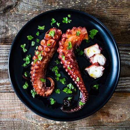 Grilled octopus tentacles with caramelized char on a black plate, garnished with fresh herbs on a rustic wooden surface